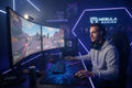 Young man at a professional blue and purple RGB gaming setup with triple monitors and mechanical keyboard